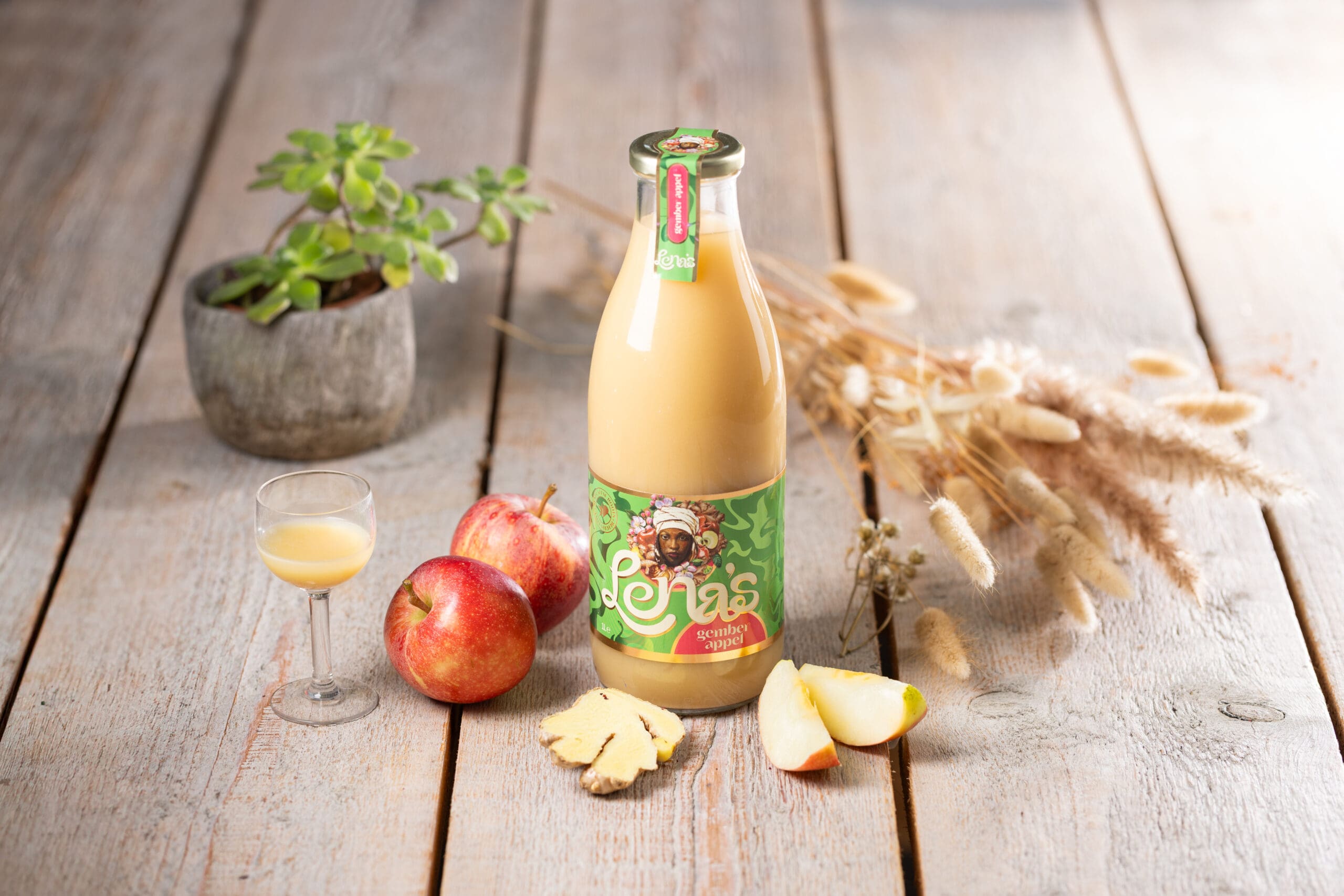 Een flesje sap van Lena's staat tussen gember- en appelpartjes op een houten tafel met plantjes op.