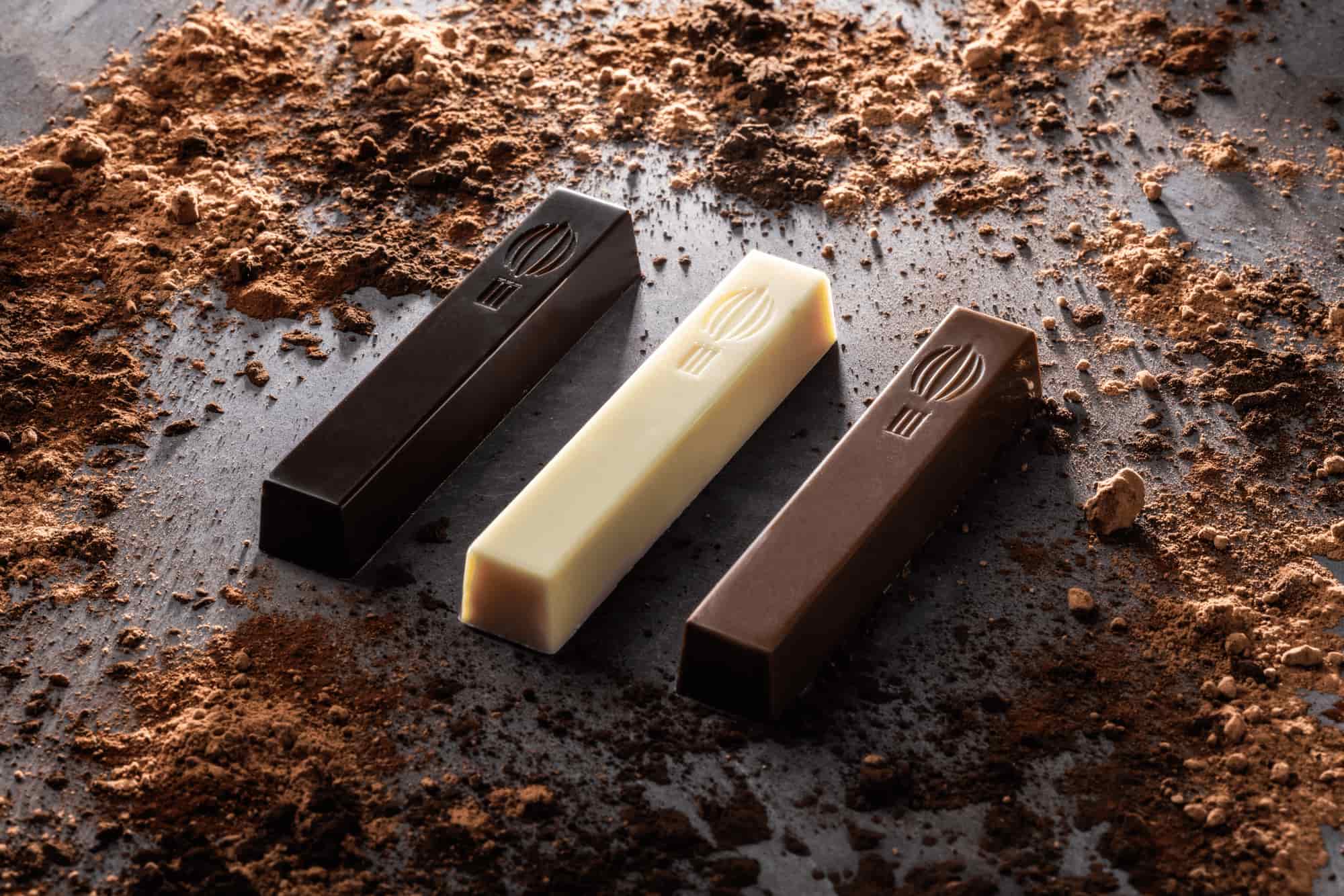 Een zwarte, bruine en witte chocoladereep liggen schuin op een grijze tafel tussen het cacaopoeder.