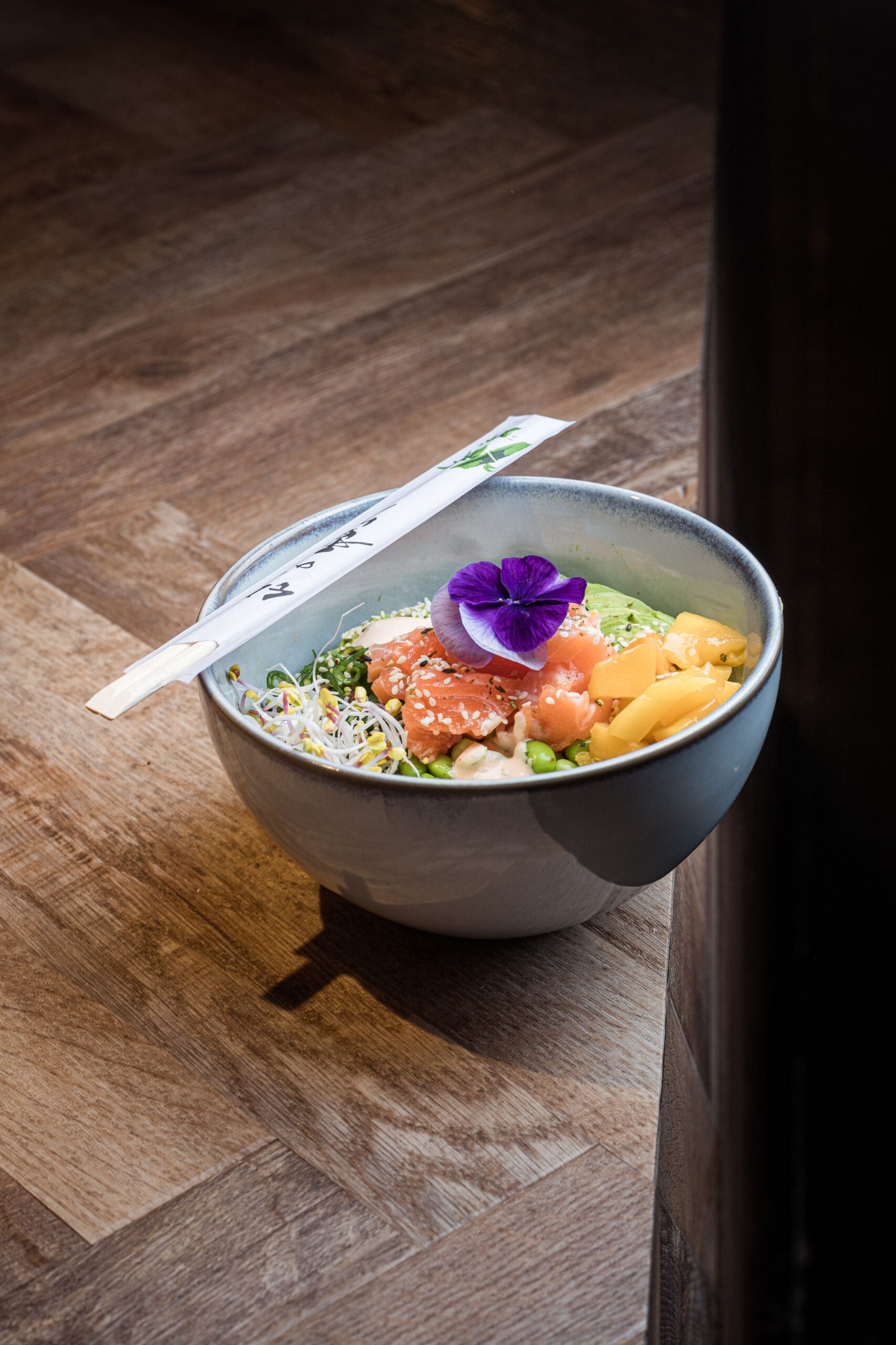 Een blauwgrijs kommetje gevuld met poké bowl, staat op de rand van een eikenhouten tafel.