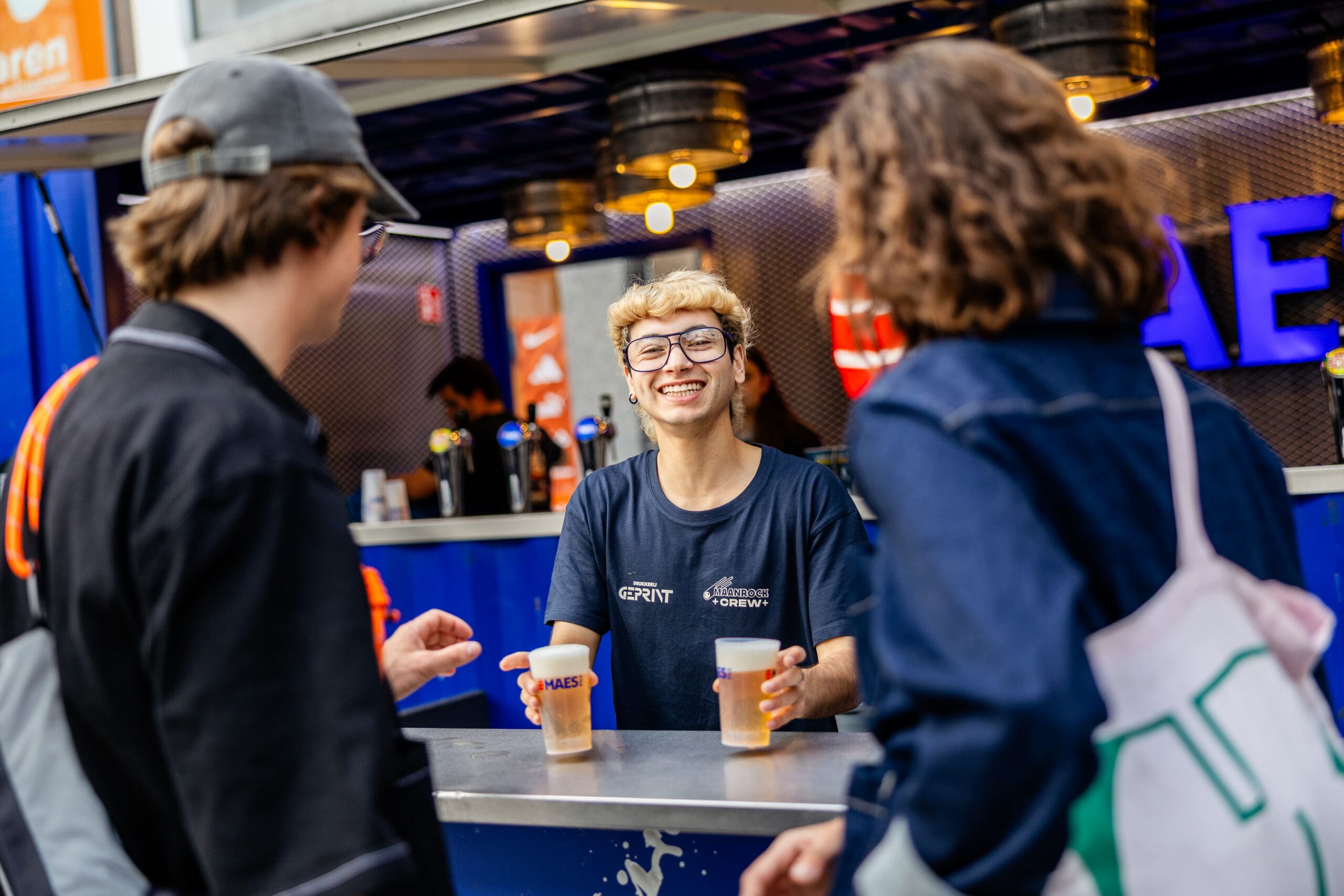 Een man achter de bar lacht naar de fotograaf terwijl hij twee biertjes afgeeft.