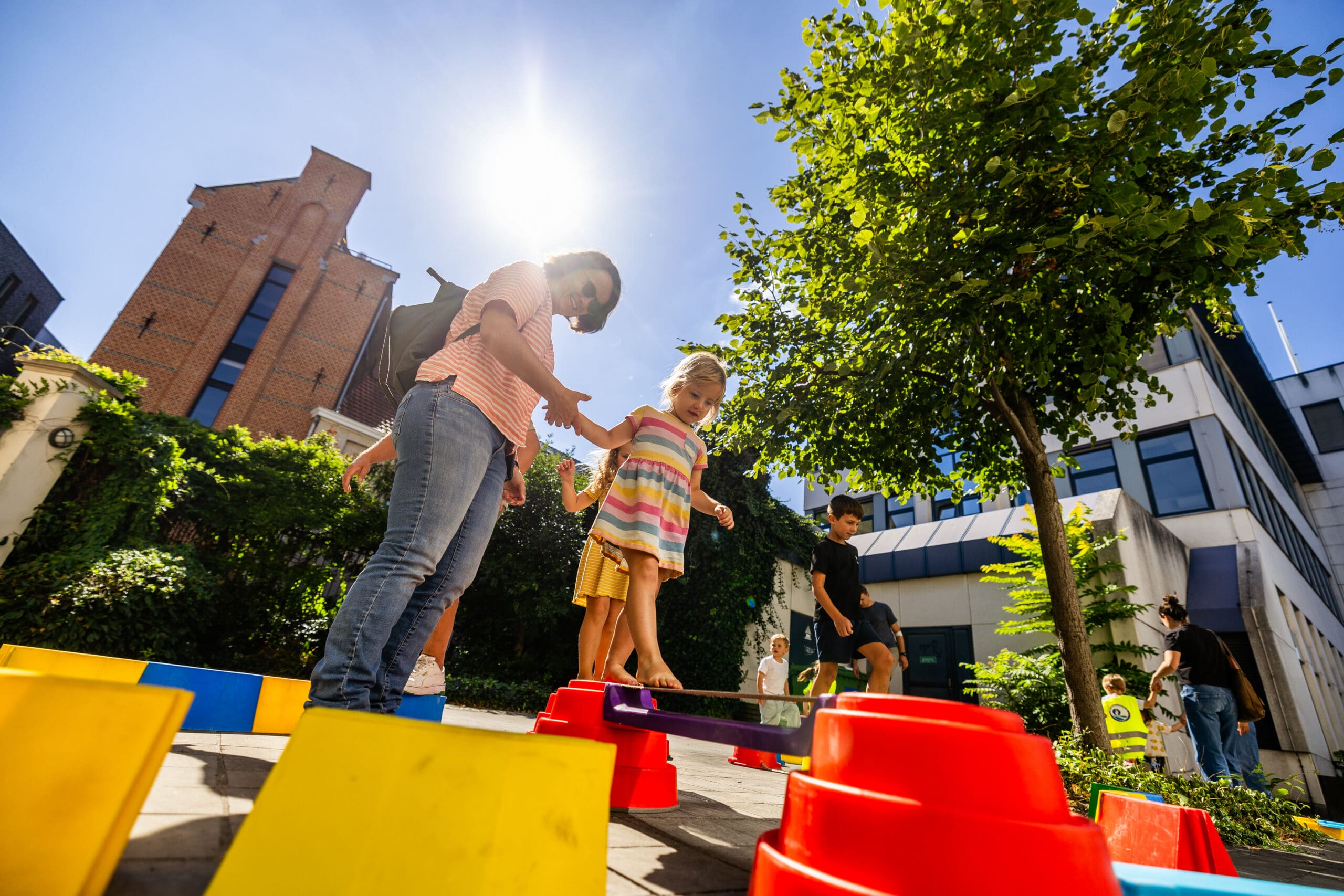 Kindje wandelt op blote voeten over een balanceerplank terwijl haar mama haar ondersteunt met een hand.